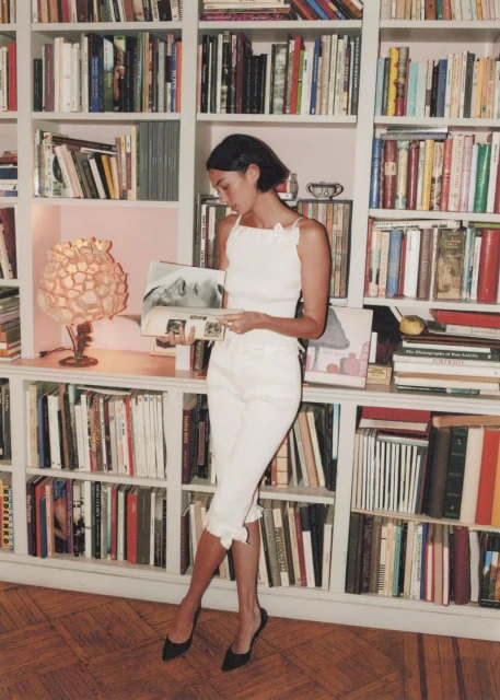 Young woman with dark hair, wearing white sleeveless top and cropped pants, stands in front of a full bookshelf in an indoor setting.