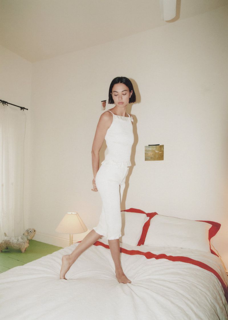 Woman in white tank top and skirt walking across a white bed in a white bedroom with lamp, artwork and a toy dog in soft light.