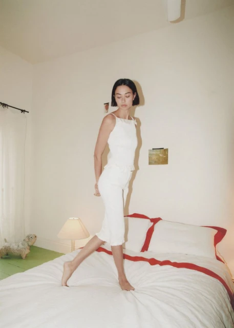 Woman in white tank top and skirt walking across a white bed in a white bedroom with lamp, artwork and a toy dog in soft light.