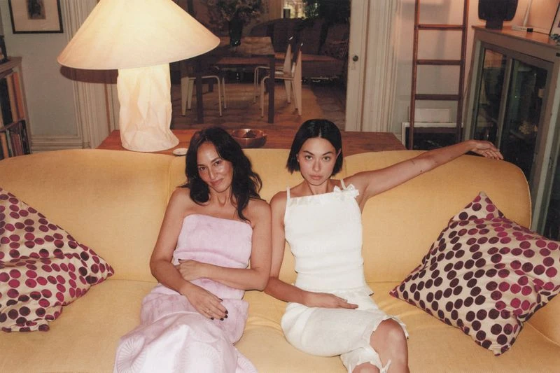 Two women sit on a yellow couch with patterned pillows in an indoor setting, illuminated by a large lamp, enjoying a relaxed, candid moment.