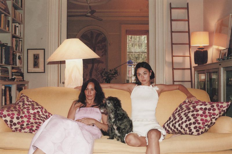 Two women and a small dog sit on a yellow sofa in an indoor setting, with a bookshelf and ladder visible in the background.