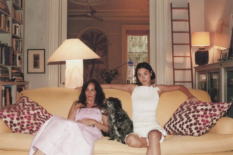 Two women and a small dog sit on a yellow sofa in an indoor setting, with a bookshelf and ladder visible in the background.