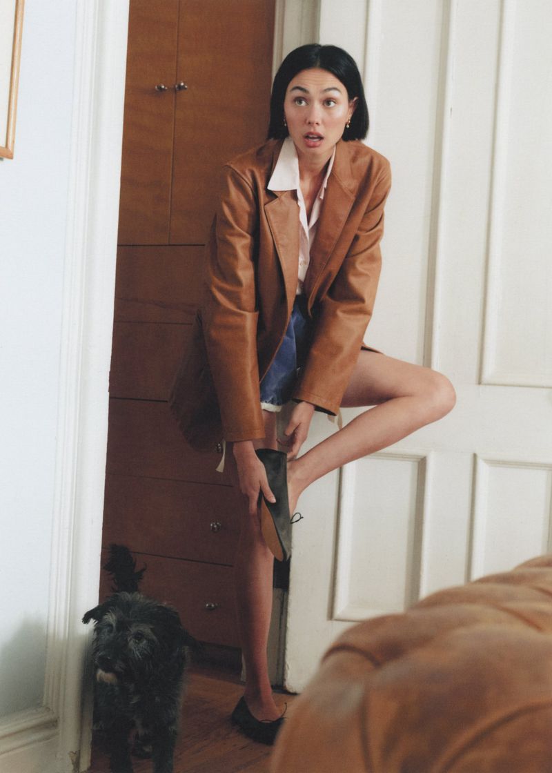 Adult woman putting on a shoe in a doorway of a home interior as a small dog looks on near a dark wood dresser.