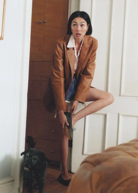 Adult woman putting on a shoe in a doorway of a home interior as a small dog looks on near a dark wood dresser.