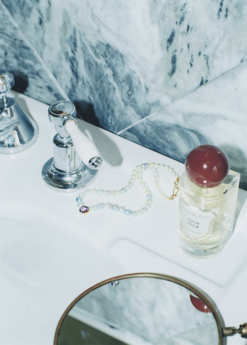 Bathroom counter with chrome faucet, glass perfume bottle with burgundy cap, pearl necklace, and vanity mirror against marble tile.