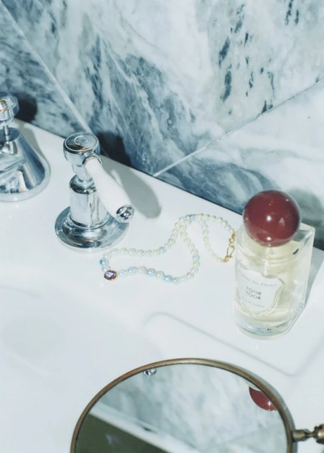 Bathroom counter with chrome faucet, glass perfume bottle with burgundy cap, pearl necklace, and vanity mirror against marble tile.