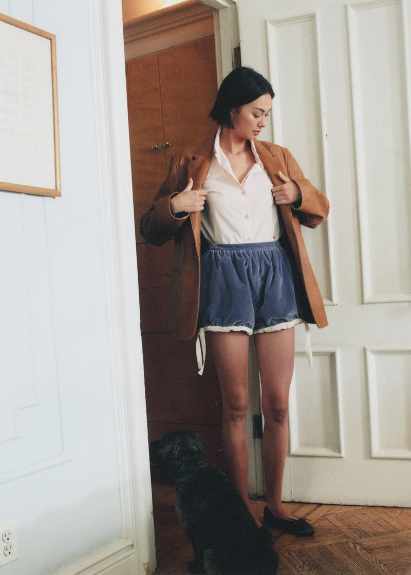 Adult woman in blazer and blue shorts standing indoors near doorway with a black dog; neutral-toned walls and a white door.