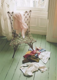 Wire frame chair with a pink draped cloth and folded garments scattered on a green wooden floor near a window.