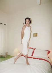 Woman with short dark hair in a white outfit standing on a white bed with red trim inside a bedroom