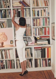 Woman in white camisole and trousers reaching up to a white bookshelf filled with books for OUTLINE Spring Catalog 2025
