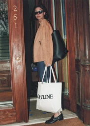 Model wearing sunglasses and a camel jacket holding a white canvas tote bag standing in a doorway