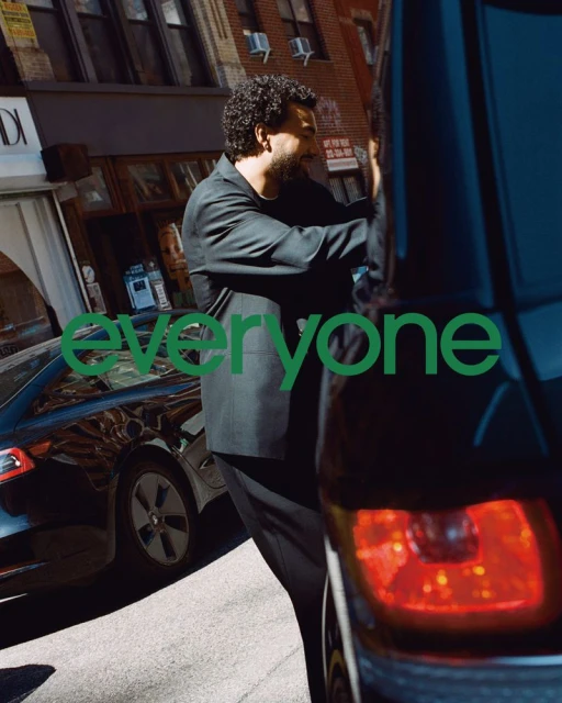 A man in a suit stands on a city street next to a dark-colored car, with commercial buildings visible and the text "everyone" overlayed.