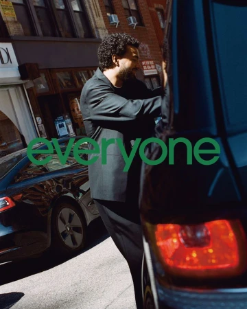 A man in a suit stands on a city street next to a dark-colored car, with commercial buildings visible and the text "everyone" overlayed.