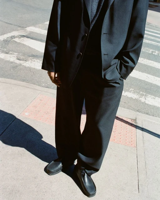 Man in a black suit and dress shoes walking on a crosswalk on a sunny day. The photo is a low angle, mid shot of the subject.