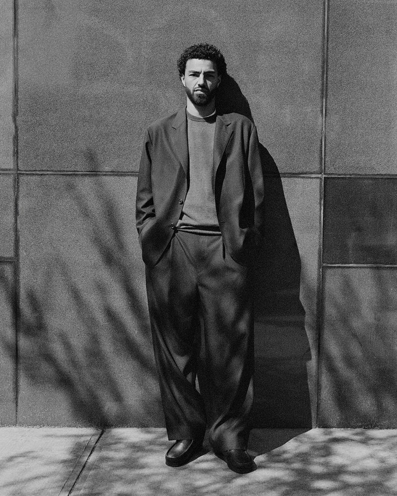 Black and white portrait of an adult male in a suit standing outdoors against a textured wall with shadow patterns from trees.