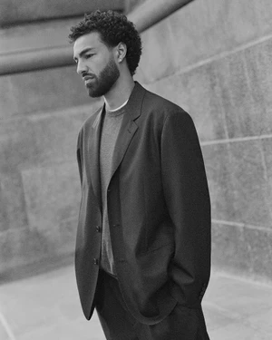Monochrome portrait of a man in a tailored blazer and crewneck sweater outdoors against stone architecture.