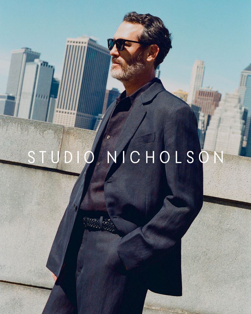 An adult male poses outdoors in a dark suit and sunglasses against a concrete barrier and cityscape background on a sunny day.