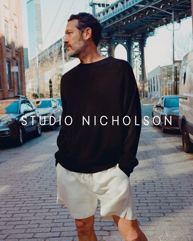Man wearing black sweater and white shorts stands on brick road near city street and bridge in daylight.