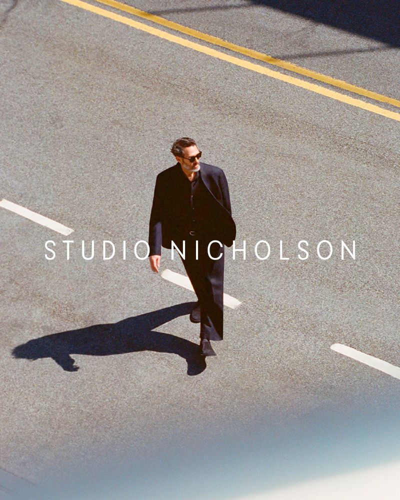 An adult male in sunglasses and a dark suit walks across an asphalt street marked with yellow and white lines, casting a long shadow.