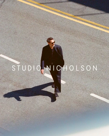 An adult male in sunglasses and a dark suit walks across an asphalt street marked with yellow and white lines, casting a long shadow.