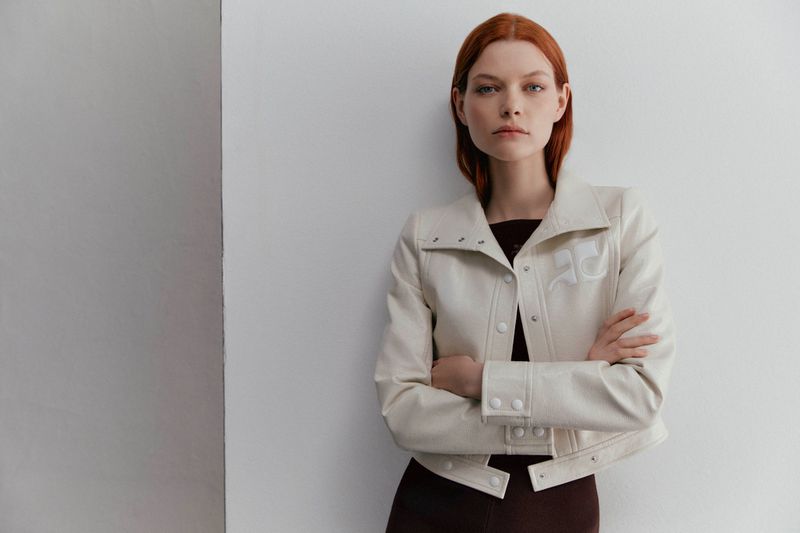 A red-haired woman poses indoors wearing an off-white jacket and a maroon dress against a two-tone white and gray wall in a portrait-style shot.