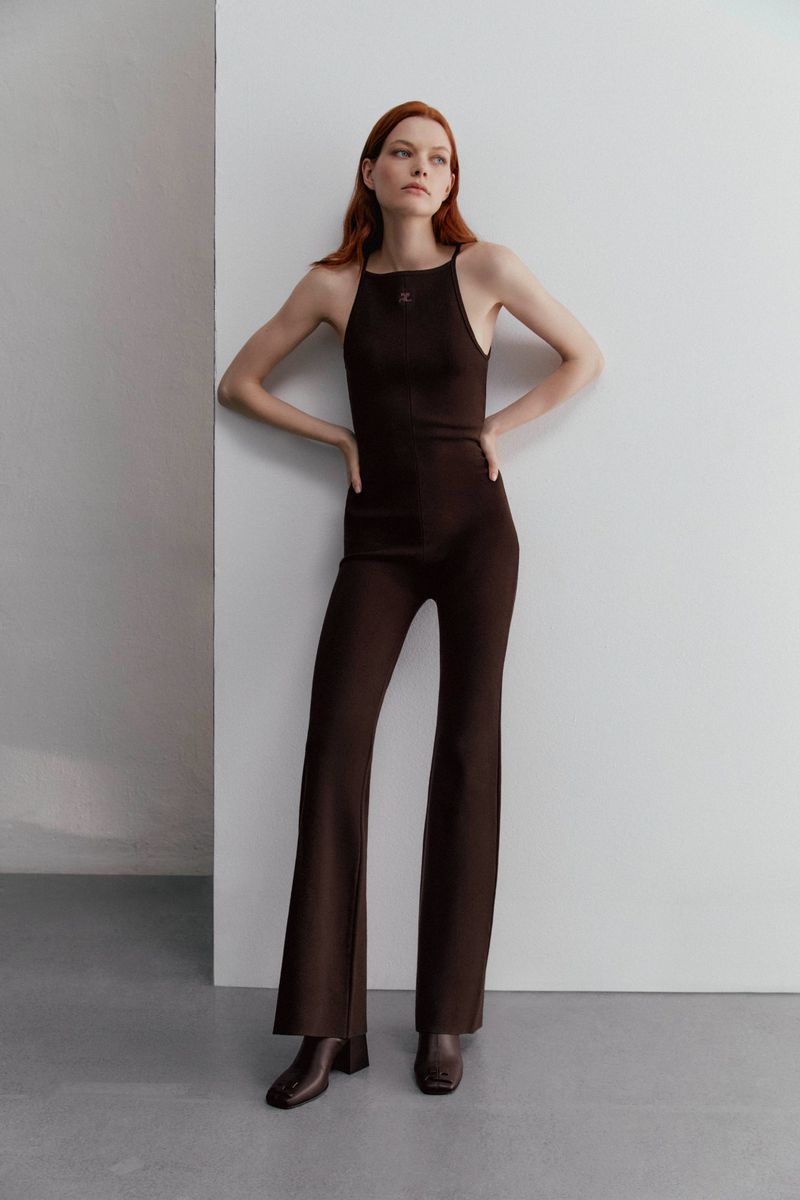 A redhead woman is posing while wearing a brown jumpsuit and brown boots in a photo studio with a white and grey background.