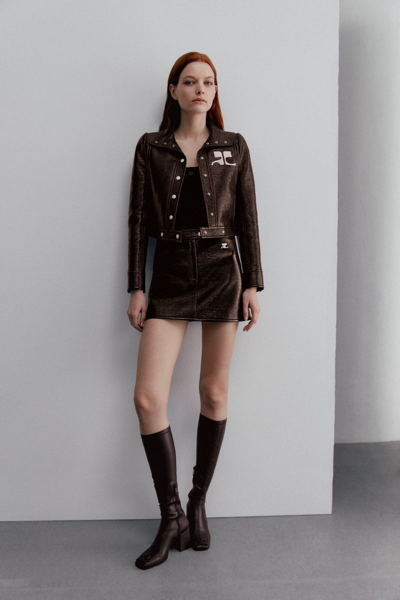Full-length shot of a woman in a dark brown textured jacket and skirt set with knee-high boots against a neutral backdrop.