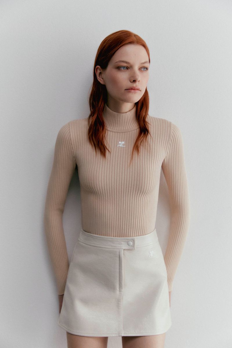 An adult woman with red hair poses in a beige turtleneck top and cream skirt against a plain white background in a studio shot.