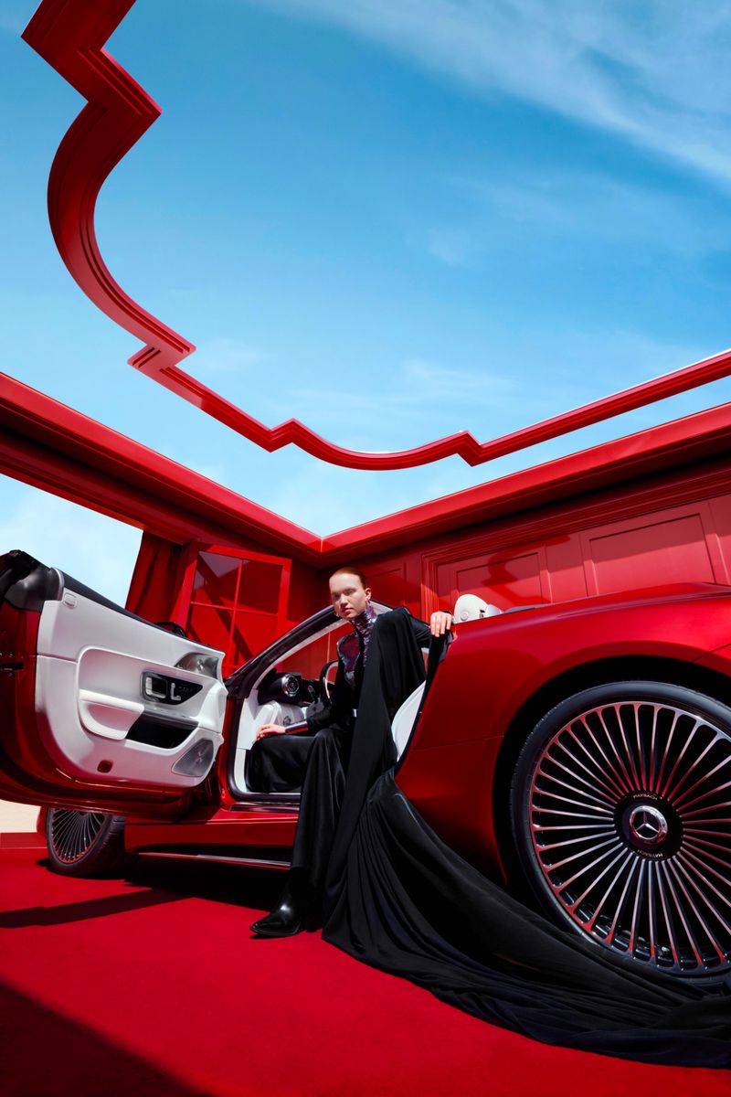 Elegant adult in a black gown poses beside a red convertible against a backdrop of red architecture and a bright blue sky.