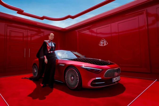 A woman in black and red stands next to a red convertible car in a matching red interior space under a blue sky visible in the background.
