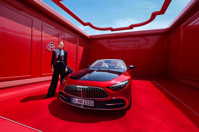 Red Maybach car in a red room with a posed adult female wearing a dark jumpsuit and silver vest in a fashion studio environment.