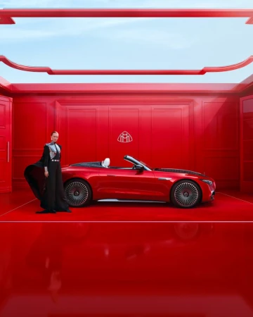 A red Maybach convertible is parked indoors with a woman in a black dress posing next to it, under bright, diffuse lighting.