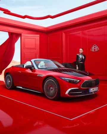 A red Maybach convertible car is parked inside an all-red room, featuring a woman standing near the wall, and a desert scene through an open doorway.