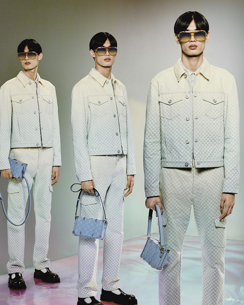 Three male models wearing patterned jackets and pants, holding bags, and standing in a photo studio with soft lighting.