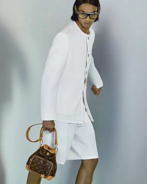 Fashion shot of a man in white suit holding a Louis Vuitton bucket bag against a light gray background. Sunglasses and short hair visible.
