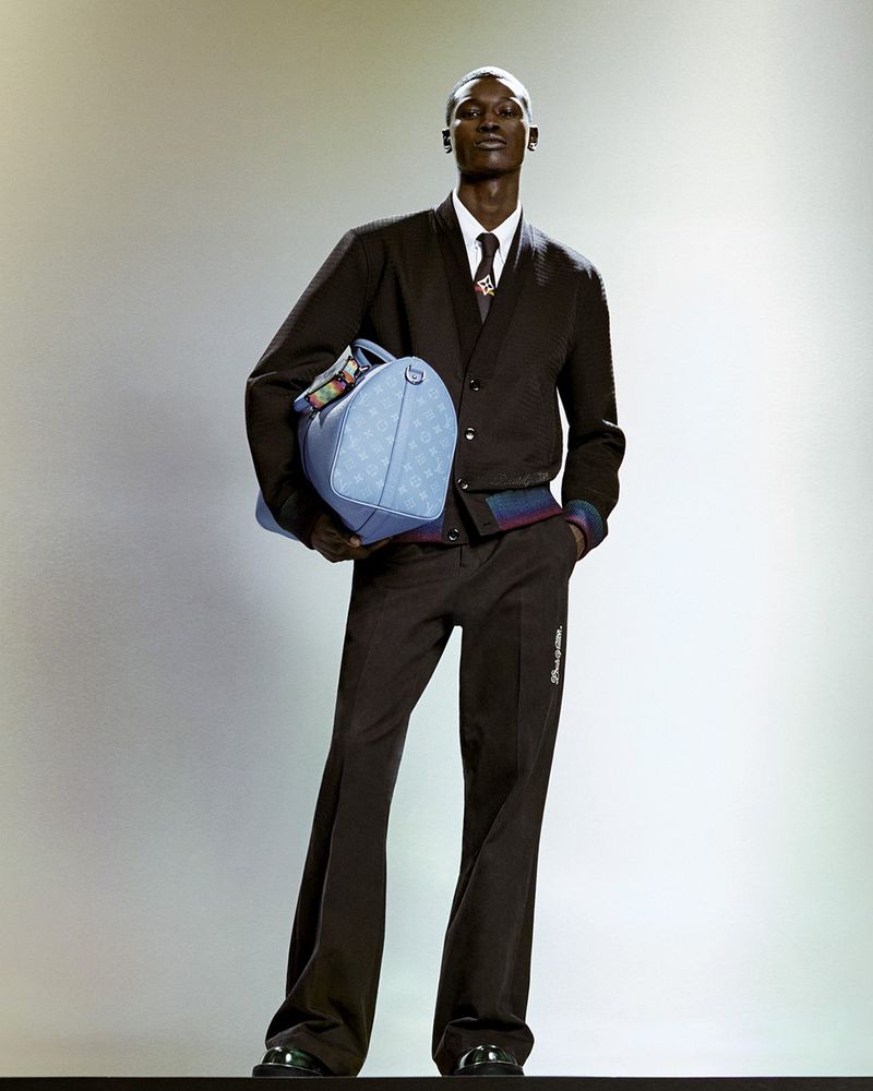 A Black male model poses in a dark brown wool suit, light blue Louis Vuitton leather duffel bag, and glossy black dress shoes against a soft gradient backdrop.