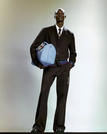 A Black male model poses in a dark brown wool suit, light blue Louis Vuitton leather duffel bag, and glossy black dress shoes against a soft gradient backdrop.