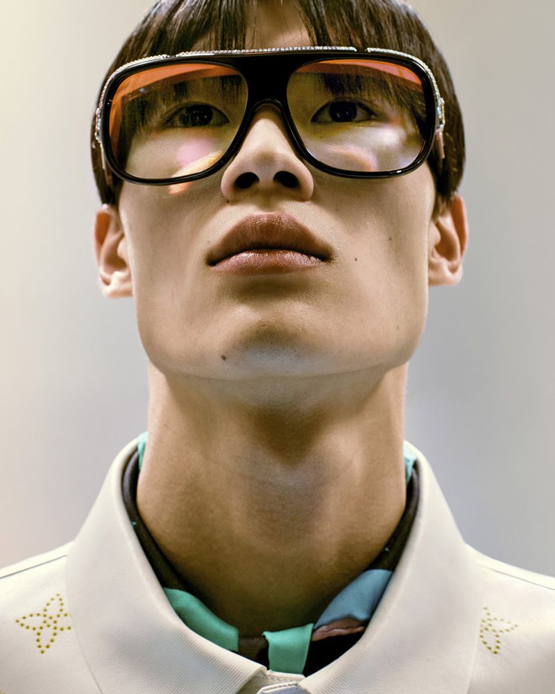 Close-up of an adult male wearing sunglasses, a collared shirt with floral embroidery, and a patterned neck scarf, shot at eye level.