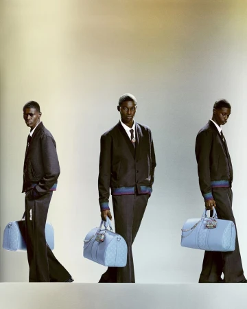 Three adult men pose with matching blue duffel bags and dark suits against a neutral gradient backdrop in a studio fashion shot.