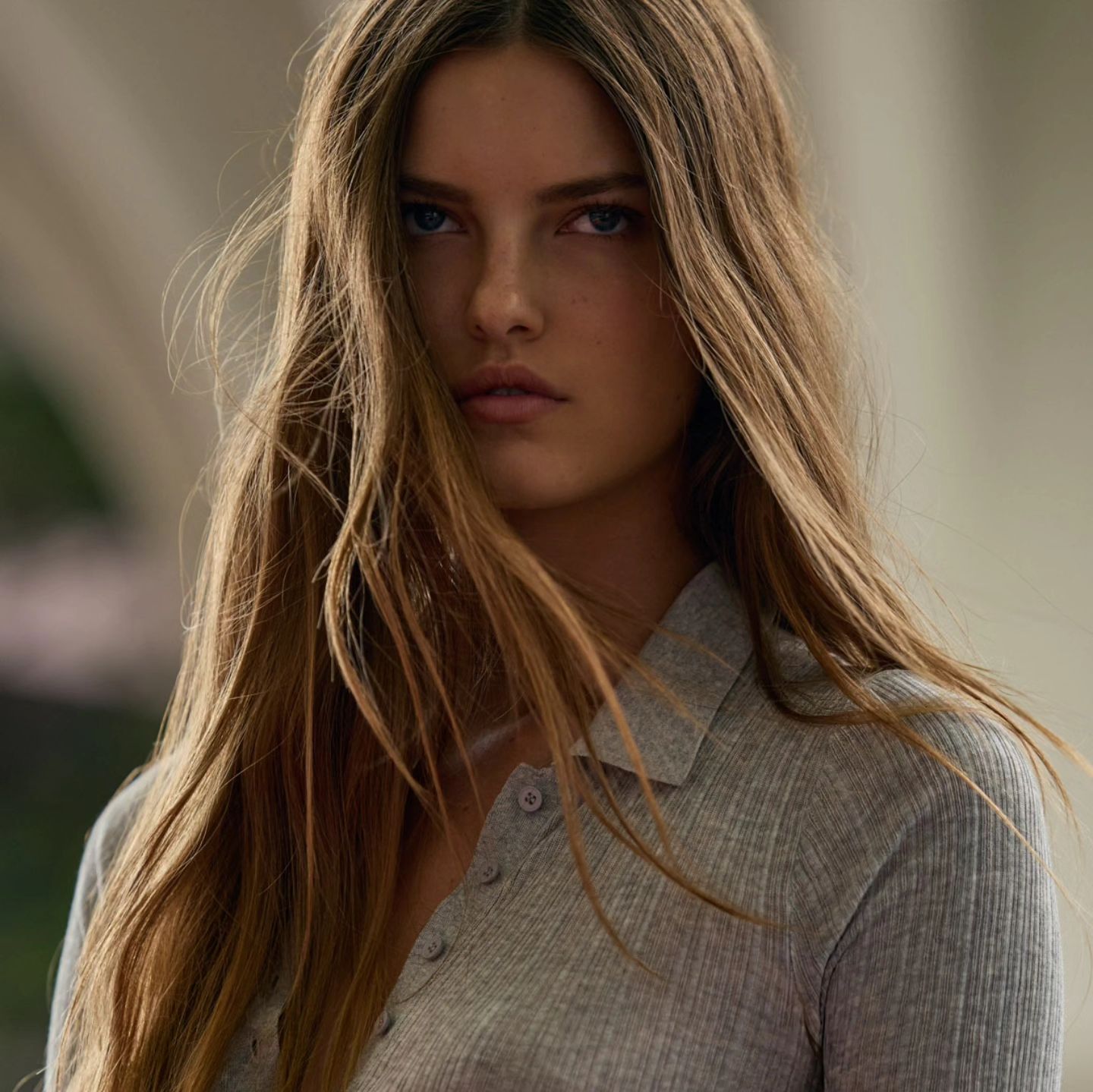 Close-up portrait of a young woman with long brown hair wearing a grey button-up shirt, looking directly at the camera with a serious expression.