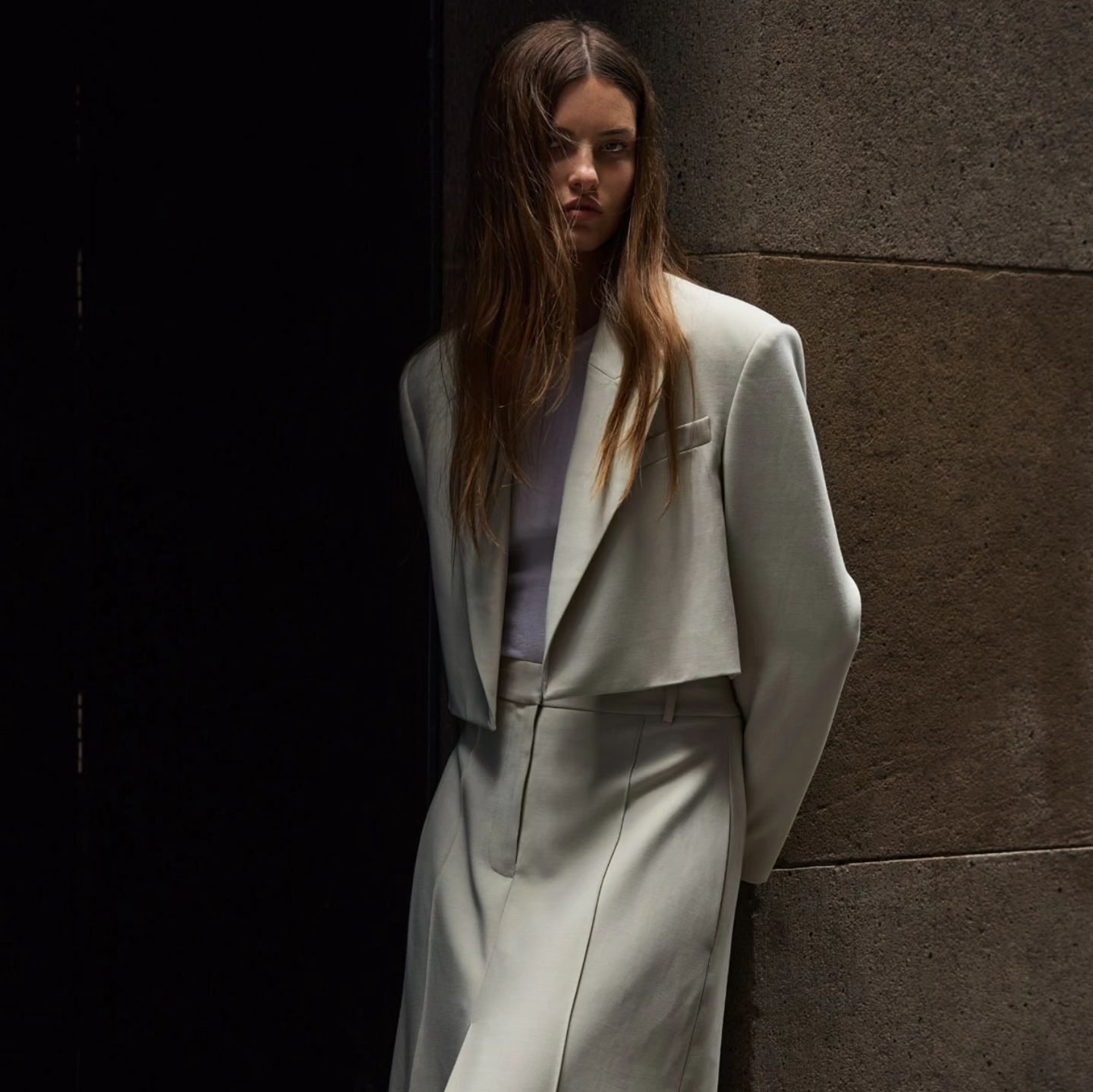 An adult female poses in a cropped light-green blazer and pants in front of a shadowed grey concrete wall, with a serious expression.