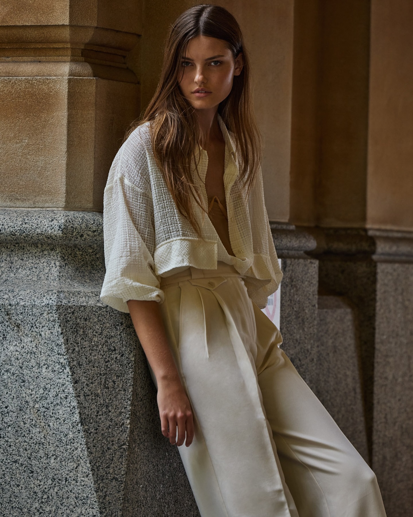 Model leaning against a stone pillar in a cream textured shirt and light trousers
