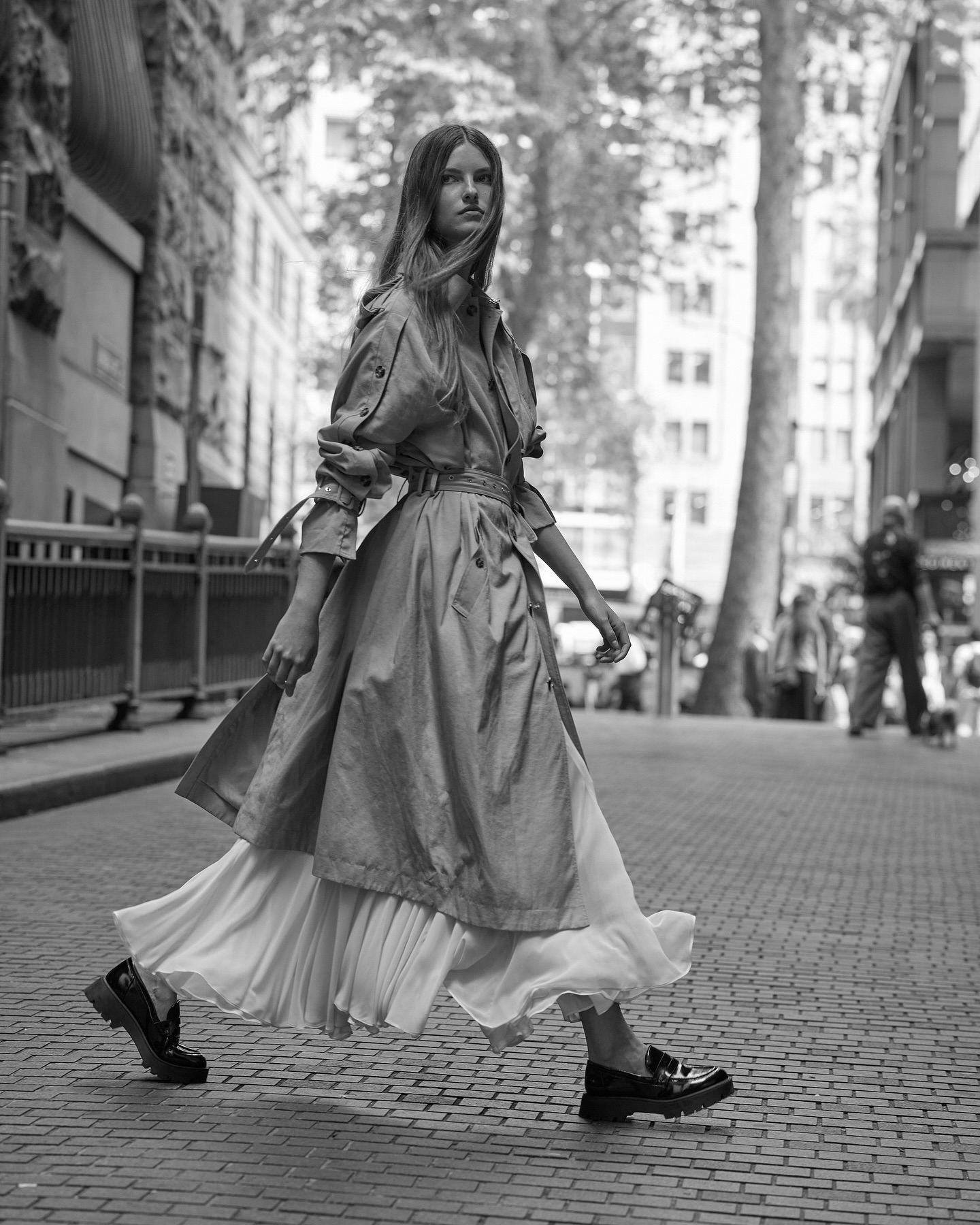 A woman walks on a city street wearing a trench coat, white dress, and black loafers. The image is black and white, creating a timeless aesthetic.