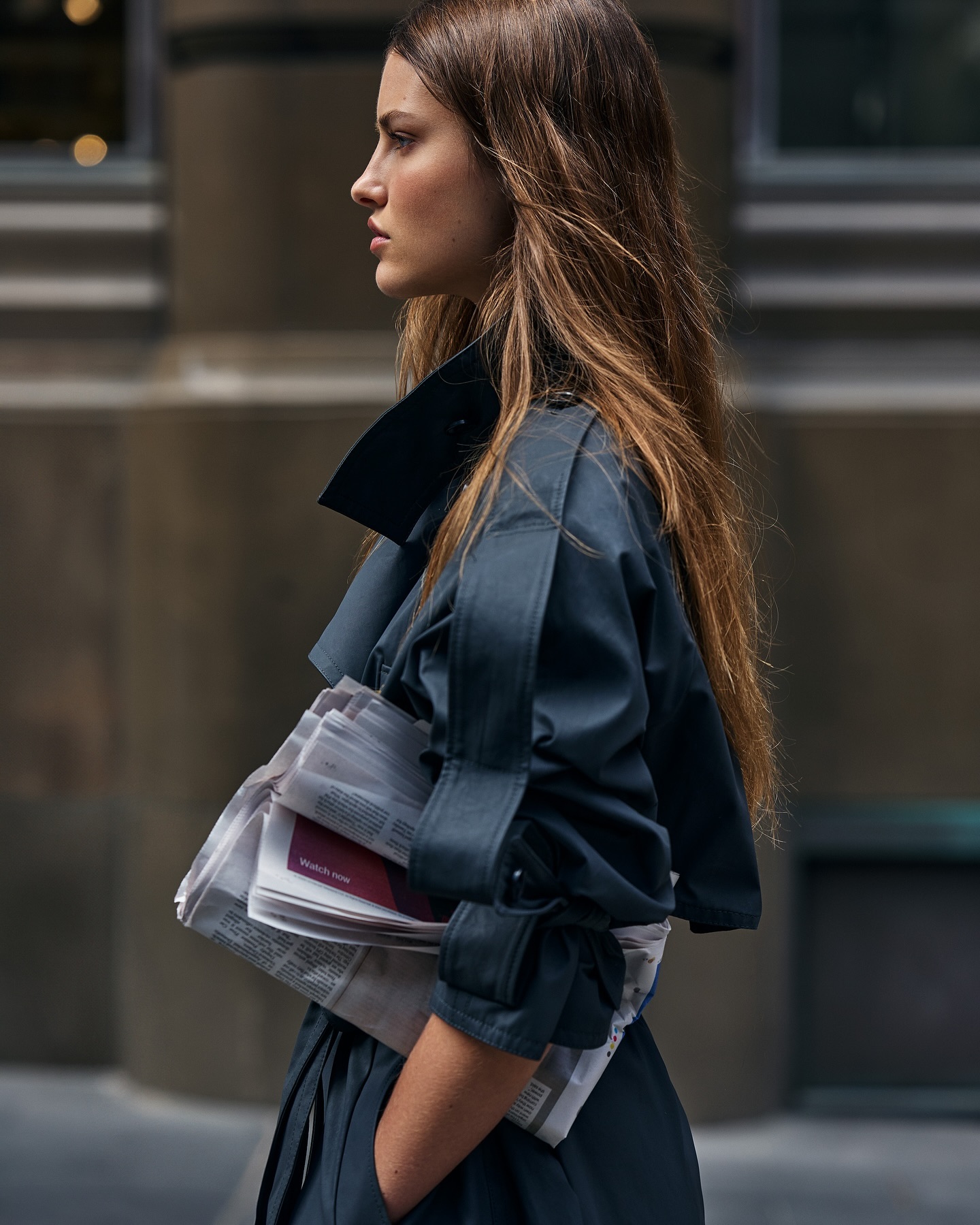 Brunette woman in blue trench coat carries newspapers on a city street. Medium shot in natural light.