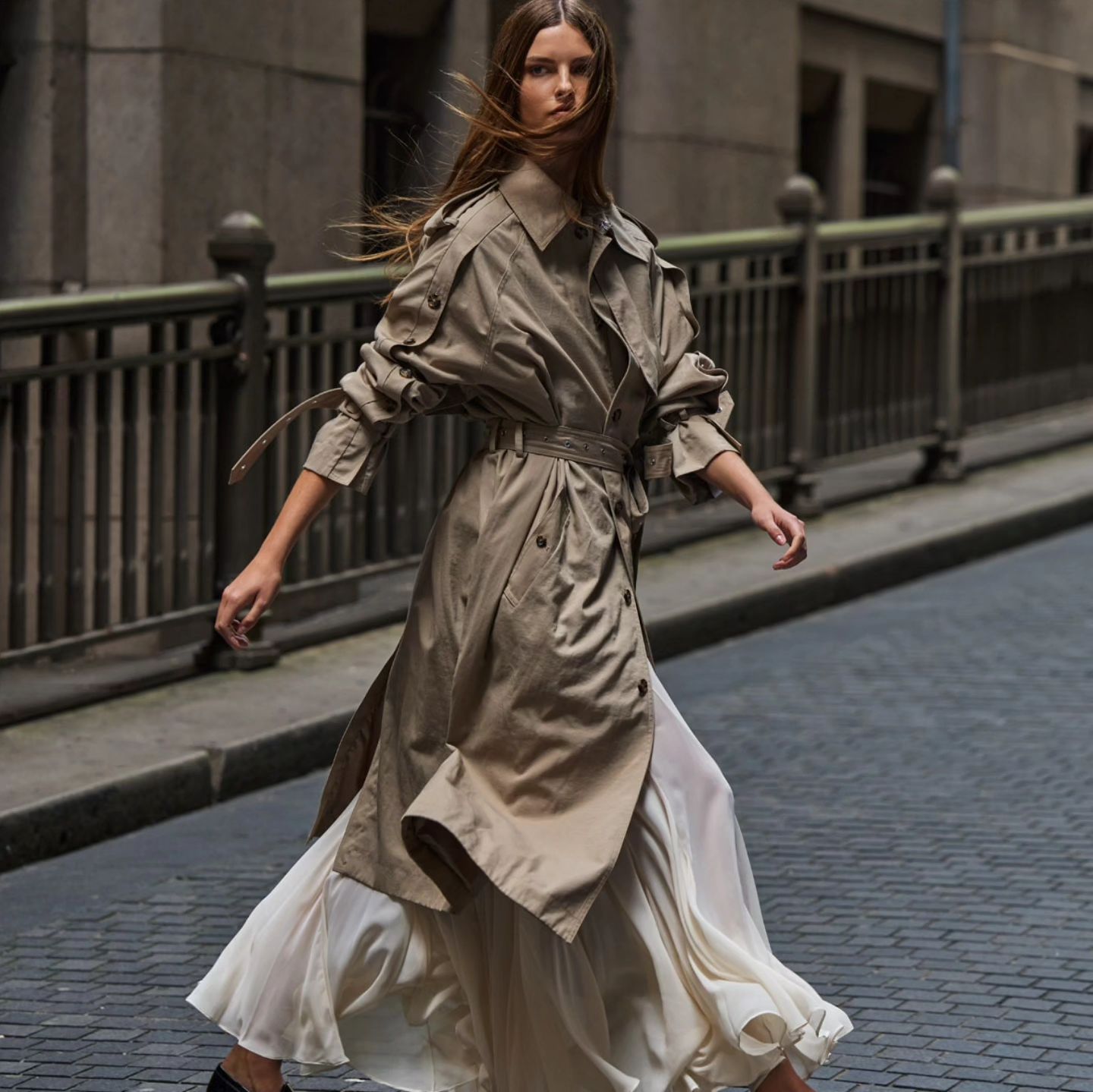 A slender woman in a trench coat and flowing skirt strides along a city street, with a neutral color palette and soft, natural lighting.