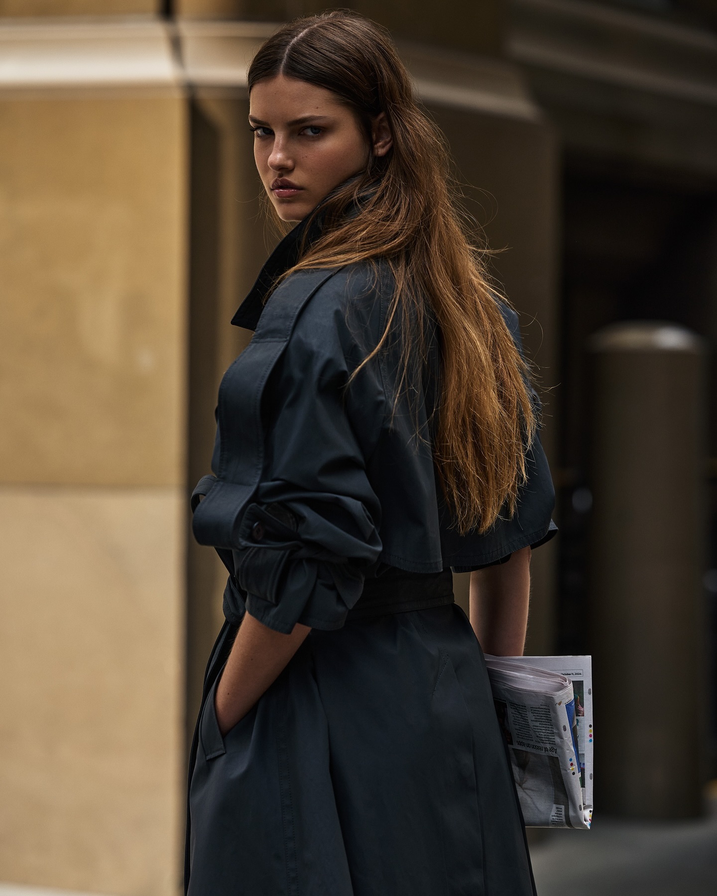 Stylish brunette woman in a black trench coat glances back over her shoulder while holding a newspaper in a city environment.