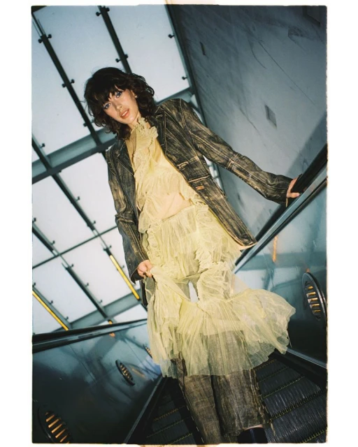 Young woman in vintage style poses on an escalator in a ruffle dress and jacket, looking at the camera from a low angle.