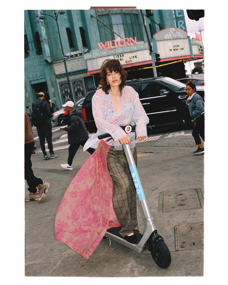 Woman rides electric scooter in the city street with a theater in the background, wearing sheer shirt and plaid pants.
