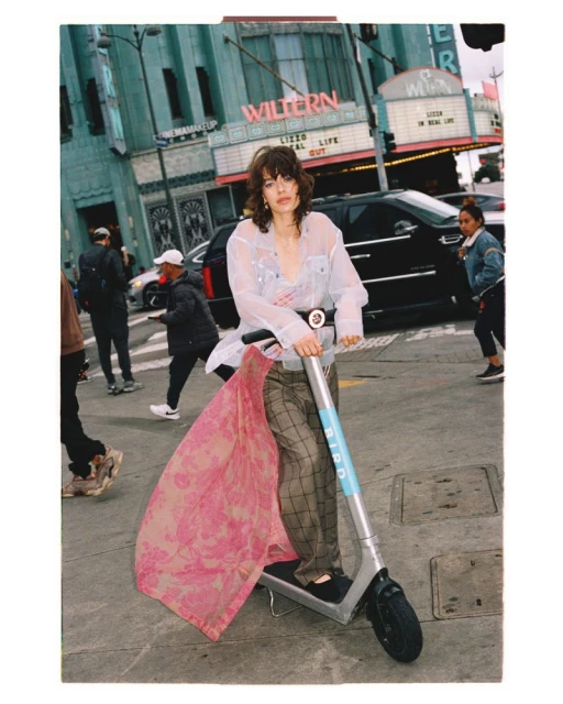 Woman rides electric scooter in the city street with a theater in the background, wearing sheer shirt and plaid pants.