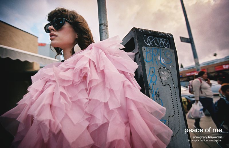 Woman in a pink ruffle coat standing next to a graffiti-covered trash can on a city street, wearing sunglasses in an editorial fashion image.